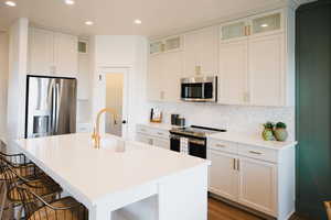 Kitchen with a center island with sink, light countertops, appliances with stainless steel finishes, glass insert cabinets, and a sink