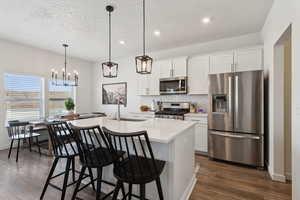 Kitchen featuring a kitchen island with sink, stainless steel appliances, light countertops, white cabinetry, and pendant lighting