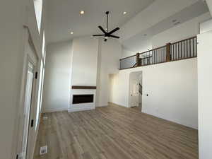 Unfurnished living room with ceiling fan, a fireplace, a towering ceiling, visible vents, and light wood finished floors