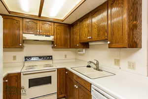 Kitchen featuring light countertops, brown cabinetry, a sink, white appliances, and under cabinet range hood