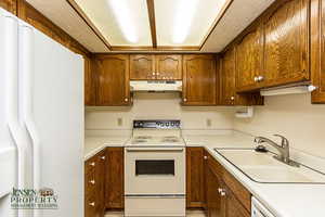 Kitchen with light countertops, brown cabinetry, a sink, white appliances, and under cabinet range hood