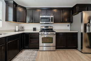 Kitchen featuring stainless steel appliances, dark brown cabinets, light countertops, and light wood-style flooring