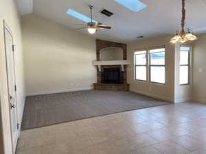 Unfurnished living room with light carpet, vaulted ceiling with skylight, visible vents, a stone fireplace, and ceiling fan with notable chandelier