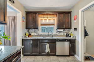 Kitchen with light stone counters, backsplash, dark brown cabinets, stainless steel dishwasher, and a sink