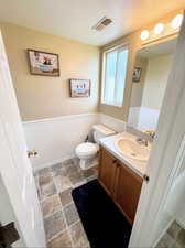 Half Bathroom featuring visible vents, toilet, stone finish floor, wainscoting, and vanity