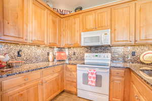 Kitchen featuring light tile patterned floors, white appliances, dark stone countertops, and tasteful backsplash