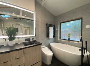 Bathroom with lofted ceiling, toilet, vanity, a freestanding tub, and tile walls