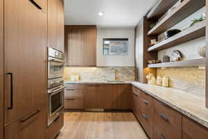 Kitchen with open shelves, backsplash, light wood-style floors, double oven, and a sink