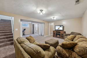Living area with a warm lit fireplace, a textured ceiling, light colored carpet, visible vents, and stairs