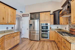 Kitchen with stainless steel appliances, light wood-style floors, custom exhaust hood, light stone countertops, and tasteful backsplash