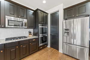 Kitchen with light stone counters, light wood-style flooring, decorative backsplash, appliances with stainless steel finishes, and dark brown cabinetry