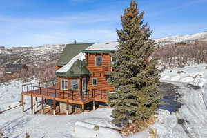 Snow covered back of property with a deck with mountain view, stone siding, and log siding