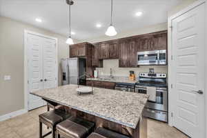 Kitchen featuring a center island with sink, stainless steel appliances, hanging light fixtures, a sink, and light stone countertops