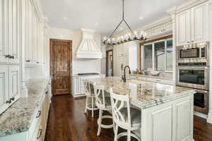 Kitchen with light stone counters, decorative light fixtures, a kitchen island with sink, custom exhaust hood, and a sink