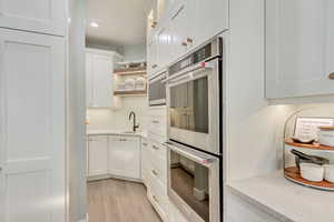 Kitchen featuring open shelves, light wood-style floors, stainless steel double oven, white cabinetry, and light stone countertops