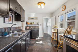 Kitchen with a sink, baseboards, dark brown cabinets, dark countertops, and stone tile flooring