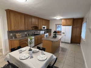 Kitchen featuring light tile patterned floors, white appliances, a kitchen island, backsplash, and brown cabinetry