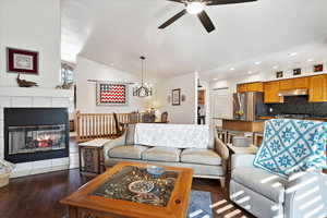 Living area with dark wood-style flooring, a fireplace, vaulted ceiling, and ceiling fan with notable chandelier