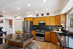 Kitchen featuring a breakfast bar area, under cabinet range hood, stainless steel appliances, a sink, and washer and dryer