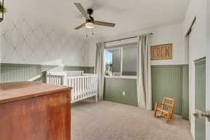 Unfurnished bedroom featuring light carpet, wallpapered walls, wainscoting, ceiling fan, and a nursery area