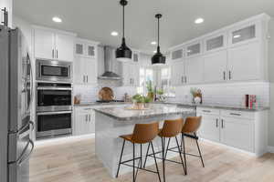 Kitchen featuring a kitchen island, appliances with stainless steel finishes, white cabinetry, light stone counters, and wall chimney exhaust hood