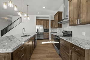 Kitchen featuring pendant lighting, appliances with stainless steel finishes, a kitchen island with sink, a sink, and wall chimney exhaust hood