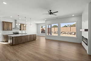 Kitchen with an island with sink, decorative light fixtures, light stone countertops, wall chimney range hood, and a sink