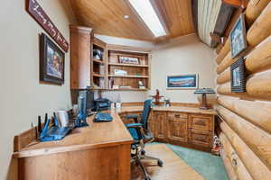 Office area featuring vaulted ceiling with skylight, light colored carpet, wood ceiling, built in study area, and rustic walls