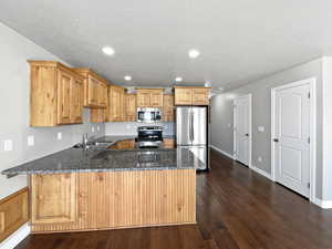 Kitchen with a peninsula, dark wood-type flooring, a sink, appliances with stainless steel finishes, and dark stone counters