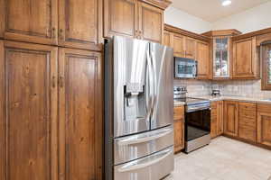 Kitchen with decorative backsplash, appliances with stainless steel finishes, brown cabinetry, glass insert cabinets, and light tile patterned flooring