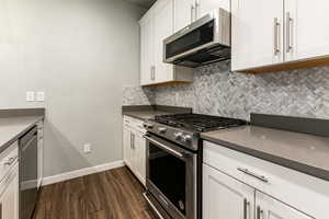 Kitchen with dark countertops, appliances with stainless steel finishes, dark wood-style flooring, white cabinetry, and backsplash