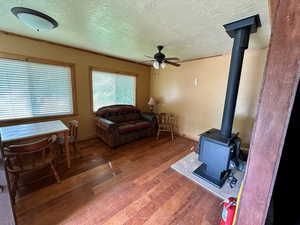 Living room with ceiling fan, a textured ceiling, baseboards, wood-type flooring, and a wood stove