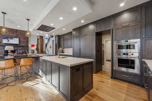 Kitchen with recessed lighting, stainless steel double oven, dark brown cabinets, light stone countertops, and light wood-type flooring