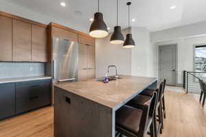 Kitchen with a sink, stainless steel built in refrigerator, modern cabinets, a center island with sink, and pendant lighting