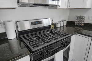 Kitchen with gas range, wall chimney exhaust hood, white cabinetry, and dark stone counters