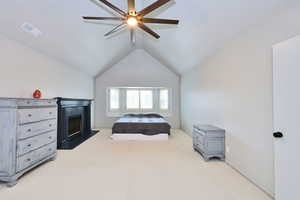 Bedroom with light carpet, visible vents, a fireplace with raised hearth, lofted ceiling, and ceiling fan