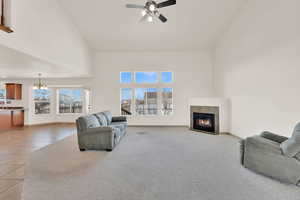 Living area with light colored carpet, a tiled fireplace, light tile patterned flooring, high vaulted ceiling, and ceiling fan with notable chandelier