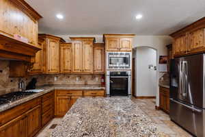 Kitchen with arched walkways, appliances with stainless steel finishes, light stone countertops, tasteful backsplash, and stone tile flooring