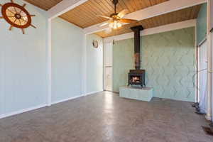 Unfurnished living room featuring a wood stove, wooden ceiling, beamed ceiling, and ceiling fan