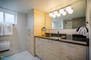 Full bathroom with a shower with shower door, vanity, baseboards, marble finish floor, and heating unit. The storage on the left of the sink has recently been replaced with a second washer/dryer.