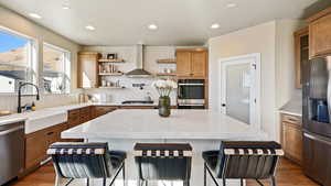 Kitchen featuring wall chimney exhaust hood, open shelves, appliances with stainless steel finishes, a sink, and a kitchen bar