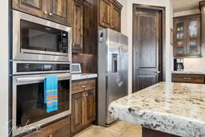 Kitchen featuring dark brown cabinetry, glass insert cabinets, light stone countertops, stainless steel appliances, and backsplash