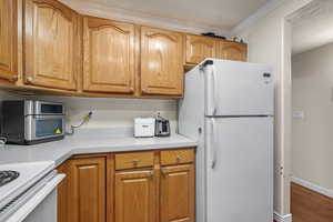 Kitchen featuring brown cabinets, crown molding, light countertops, white appliances, and baseboards