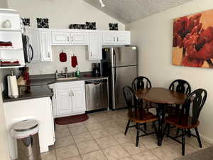 Kitchen featuring stainless steel appliances, dark countertops, lofted ceiling, white cabinets, and a sink