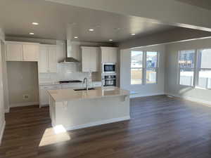 Kitchen featuring a kitchen island with sink, a sink, white cabinetry, appliances with stainless steel finishes, and wall chimney exhaust hood