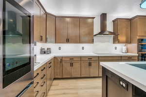 Kitchen featuring stainless steel double oven, black electric cooktop, light countertops, wall chimney exhaust hood, and light wood finished floors