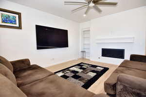 Living room with built in features, baseboards, a ceiling fan, a glass covered fireplace, and wood finished floors