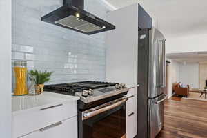 Kitchen featuring dark wood finished floors, tasteful backsplash, appliances with stainless steel finishes, white cabinets, and wall chimney range hood