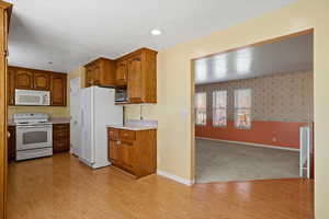 Kitchen with brown cabinetry, light wood-type flooring, white appliances, and baseboards