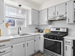 Kitchen with decorative backsplash, white cabinets, stainless steel gas stove, a sink, and under cabinet range hood